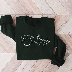 May include: A dark green sweatshirt with a white graphic design featuring a sun and a moon with the text "Live by the sun, love by the moon."