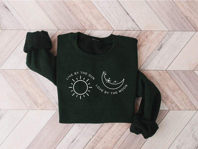 May include: A dark green sweatshirt with a white graphic design featuring a sun and a moon with the text "Live by the sun, love by the moon."