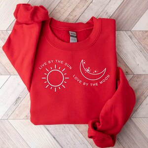 May include: Red sweatshirt with a white graphic design that says "Live by the sun, love by the moon" with a sun and moon illustration.