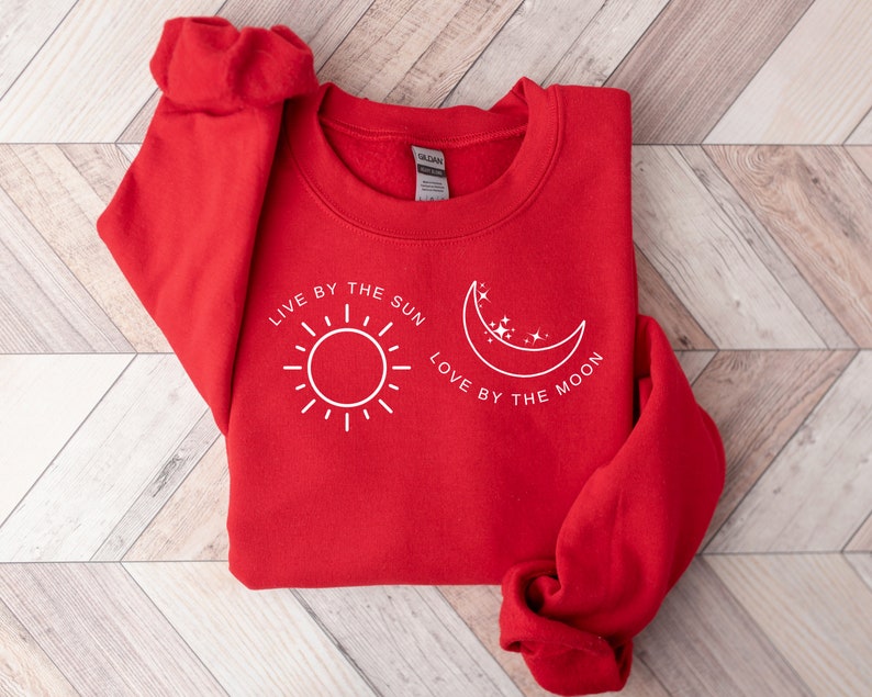 May include: Red sweatshirt with a white graphic design that says "Live by the sun, love by the moon" with a sun and moon illustration.