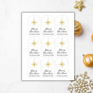 Puede incluir: Hoja de etiquetas de regalo navideñas blancas con un diseño de estrella dorada y las palabras "Merry Christmas" en escritura negra. Las etiquetas también incluyen el texto "The Johnson Family". Adornos y decoraciones navideñas doradas rodean las etiquetas.