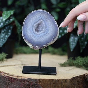 May include: A purple and white agate slice on a black metal stand. The agate has a natural crystal formation in the center.
