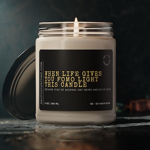 Puede incluir: Una vela blanca en un tarro de vidrio con una etiqueta negra que dice "WHEN LIFE GIVES YOU FOMO LIGHT THIS CANDLE BECAUSE FEAR OF MISSING OUT NEVER SMELLED SO GOOD." La etiqueta también incluye "9 OZ 266 ML" y "50-60 HOUR BURN".