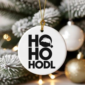 Puede incluir: Un adorno navideño de cerámica blanca con el texto "HO HO HODL" en negro y un dibujo de gorro de Papá Noel. El adorno cuelga de un cordón dorado, con otros adornos y un árbol de Navidad nevado en el fondo.