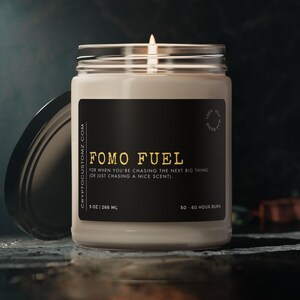 Puede incluir: Un tarro de vela negro y dorado con el texto "FOMO FUEL" y el lema "Para cuando persigues la próxima gran cosa (o simplemente persigues un buen aroma)." El tarro de vela es de 9 oz (266 ml) y tiene un tiempo de combustión de 50 a 60 horas.