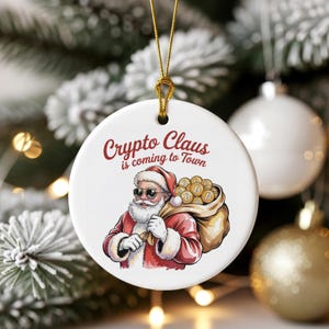 Puede incluir: Adorno navideño redondo blanco con una cuerda dorada. Presenta una ilustración de Papá Noel con gafas de sol y llevando un saco lleno de símbolos Bitcoin. El texto "Crypto Claus is coming to Town" está escrito en rojo.