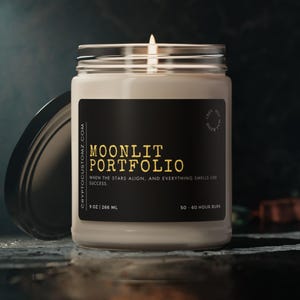 May include: A black and gold candle jar with the text "MOONLIT PORTFOLIO" and the tagline "WHEN THE STARS ALIGN, AND EVERYTHING SMELLS LIKE SUCCESS." The jar is 9 oz (266 ml) and has a 50-60 hour burn time.