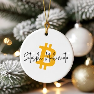 Puede incluir: Adorno navideño redondo blanco con un símbolo Bitcoin dorado y las palabras "Satoshi Nakamoto" en escritura negra. El adorno cuelga de una cuerda dorada. Otras decoraciones navideñas y vegetación están en el fondo.