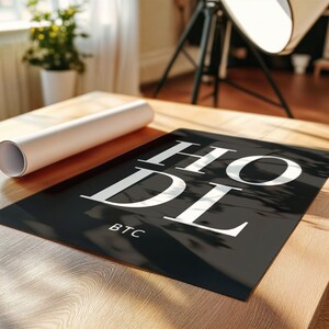 May include: Black and white poster with the word "HODL" in large white letters and "BTC" in smaller white letters below. The poster is on a wooden surface.