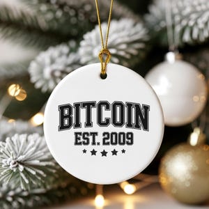 Puede incluir: Adorno navideño blanco y redondo con la palabra "BITCOIN" en letras negras. Debajo, "EST. 2009" y cinco estrellas negras. El adorno cuelga de un cordón dorado.