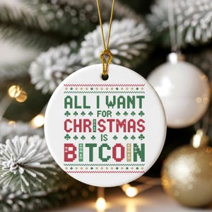 Puede incluir: Adorno navideño redondo y blanco con el texto "ALL I WANT FOR CHRISTMAS IS BITCOIN" en verde y rojo. El adorno tiene una cuerda dorada para colgar y un patrón festivo.