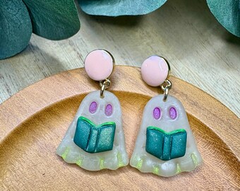 May include: A pair of handmade ghost earrings. Each earring features a light pink stud, a translucent ghost shape with purple eyes, and a teal book. The earrings are on a wooden surface.