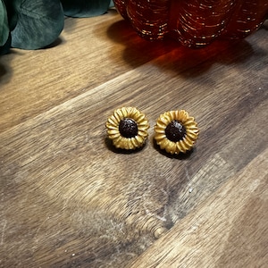 Puede incluir: Un par de pendientes de girasol dorados con centros marrones. Los pendientes están sobre una superficie de madera.