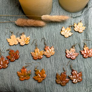 May include: A set of 10 pairs of ceramic earrings shaped like maple leaves. The leaves are painted in shades of brown, orange, and yellow, resembling autumn foliage.