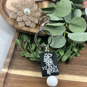 May include: Black rectangular keychain with the text "In My Villain Era" in white. The keychain has a silver ring, a white bead, and a black bead. The background includes greenery and a wooden tray.