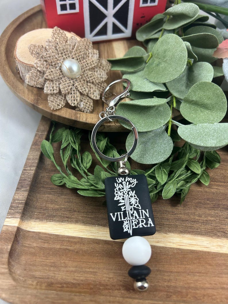 May include: Black rectangular keychain with the text "In My Villain Era" in white. The keychain has a silver ring, a white bead, and a black bead. The background includes greenery and a wooden tray.