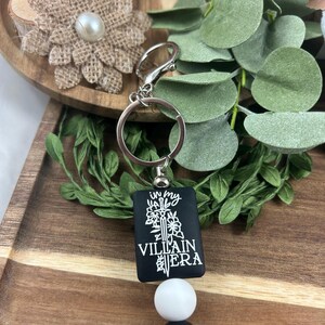 May include: A black keychain with the text "in my VILLAIN ERA" in white. The keychain has a silver ring and clasp, a white bead, and black and silver accent beads. The background includes greenery and a wooden tray.