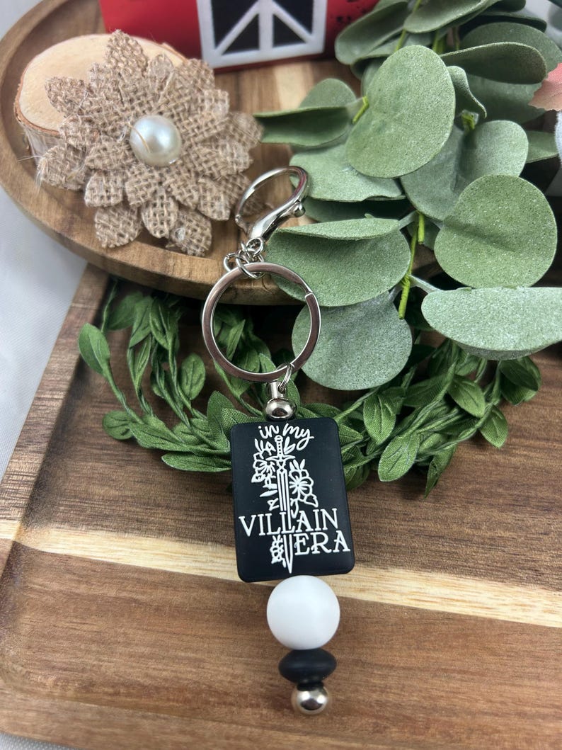 May include: A black keychain with the text "in my VILLAIN ERA" in white. The keychain has a silver ring and clasp, a white bead, and black and silver accent beads. The background includes greenery and a wooden tray.