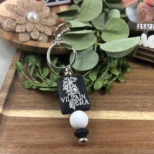 May include: A black keychain with the text "In my Villain Era" in white. The keychain has a silver ring and a white and black bead accent. The background includes greenery and a wooden tray.