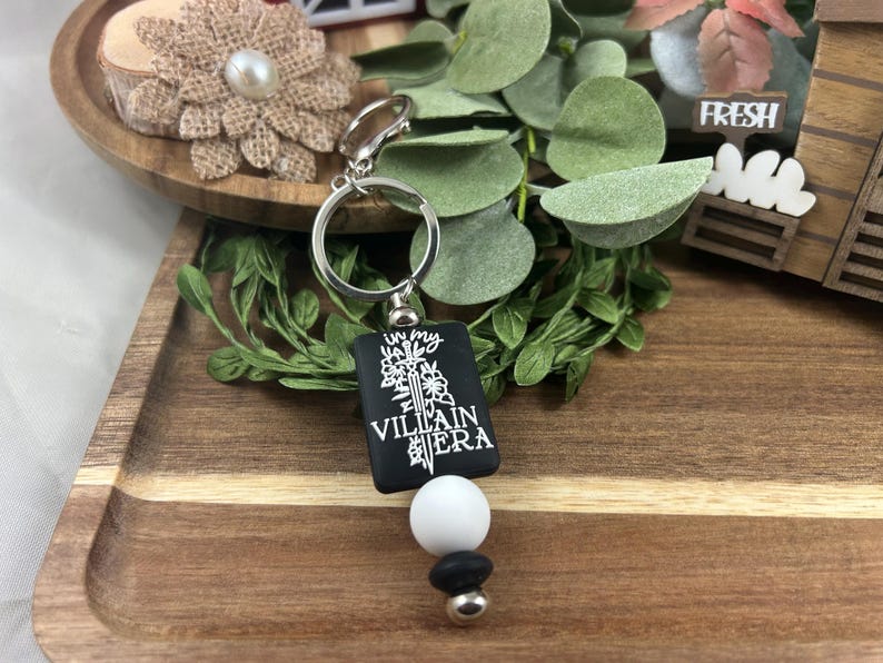 May include: A black keychain with the text "In my Villain Era" in white. The keychain has a silver ring and a white and black bead accent. The background includes greenery and a wooden tray.