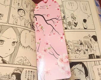 May include: A pink bookmark with a cherry blossom design. The bookmark features a branch with pink flowers and a small hole at the top. The bookmark is placed on top of a manga comic book.
