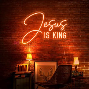 May include: A neon sign with the words "Jesus is King" in a cursive font. The sign is lit up in a warm orange color and is mounted on a brick wall.