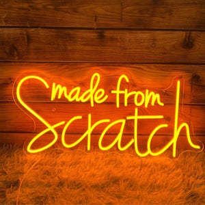 May include: A neon sign in yellow script reads "made from scratch" against a wooden background. The sign is outlined in a clear acrylic backing and sits on a bed of orange fur.