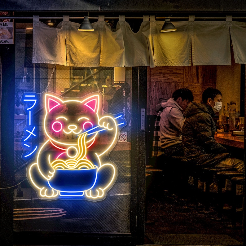 Puede incluir: Un letrero de ne&oacute;n de un gato rosa y amarillo comiendo fideos ramen. El letrero est&aacute; en la ventana de un restaurante con dos personas sentadas en una mesa en el fondo.