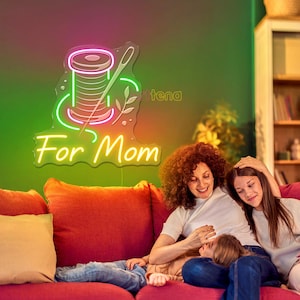 Op de afbeelding: Een neonbord met een garenklos en een naald, met de woorden "For Mom" in een cursief lettertype. Het bord is verlicht met roze, groene en gele lichten. Het is gemonteerd op een groene muur. Een moeder en twee kinderen zitten op een rode bank.
