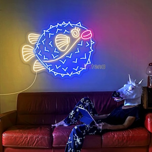 May include: A neon sign shaped like a pufferfish, with blue, white, and pink lights, hangs on a wall. The sign is above a red leather couch. A person wearing a unicorn mask is sitting on the couch, holding a tablet.