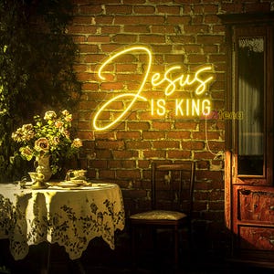 May include: A yellow neon sign with the words "Jesus is King" on a brick wall background. The sign is hanging on a wall in a room with a table and chair.
