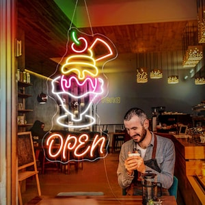 Könnte beinhalten: Ein Neon-Schild in einem Café-Schaufenster zeigt ein farbenfrohes Eisbecher-Design mit dem Wort "Open" in Schreibschrift. Das Schild wird von hellroten, gelben und weißen Lichtern beleuchtet und hängt vor einem Café-Interieur.