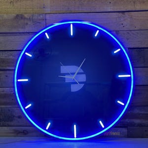 May include: A round, black wall clock with a bright blue neon light border. The clock face features a white Dash symbol and white hands. The clock has white markers for the hours. The clock is mounted on a wooden wall.