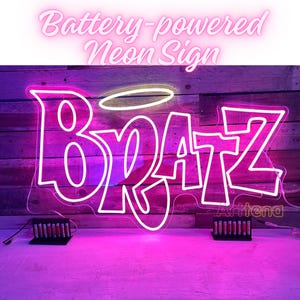 以下が含まれることがあります： ピンクと白のバッテリー駆動ネオンサインで、「BRATZ」と「Z」の上にハローがあります。サインは木製の背景にあり、上には「Battery-powered Neon Sign」という文字があります。サインは明るいピンク色に光っています。