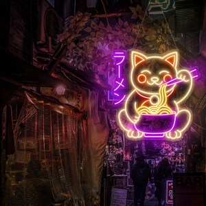 Puede incluir: Letrero de ne&oacute;n de un gato de dibujos animados sosteniendo un taz&oacute;n de fideos ramen con palillos. El letrero est&aacute; iluminado en rosa y amarillo y dice "Ramen" en caracteres japoneses.
