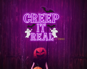 Op de afbeelding: Een wit neonbord met de tekst "Creep It Real", met vleermuizen en geesten, tegen een paarse houten achtergrond. Een persoon houdt een gesneden pompoenhoofd en een gesneden pompoen vast.
