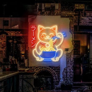 Puede incluir: Un letrero de ne&oacute;n de un gato de dibujos animados comiendo fideos ramen con la palabra japonesa "Ramen" escrita en rojo y blanco en el lateral. El gato es amarillo con un collar rojo y sostiene palillos en su pata derecha.