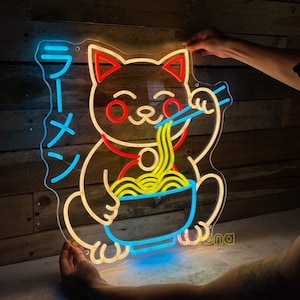 Puede incluir: Se&ntilde;al de ne&oacute;n de un gato de dibujos animados comiendo fideos ramen. El gato es amarillo con orejas rojas y un collar rojo. Los ramen son amarillos y el taz&oacute;n es azul. El letrero dice "Ramen" en caracteres japoneses.