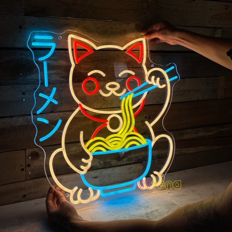 Puede incluir: Se&ntilde;al de ne&oacute;n de un gato de dibujos animados comiendo fideos ramen. El gato es amarillo con orejas rojas y un collar rojo. Los ramen son amarillos y el taz&oacute;n es azul. El letrero dice "Ramen" en caracteres japoneses.