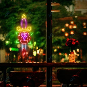 May include: A neon sign depicting a dagger with a rose wrapped around the blade. The sign is in a window with a blurry background of a cafe with lights and a vase of red roses.