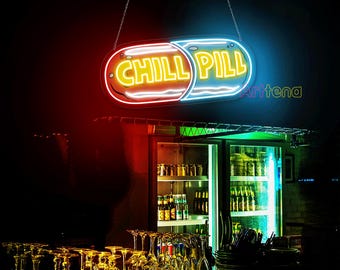 May include: A neon sign shaped like a pill with the words "Chill Pill" in yellow letters on a blue and red background. The sign is hanging above a bar with a refrigerator behind it.