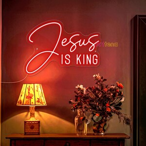 May include: Red neon sign with the words "Jesus is King" on a red wall with a lamp and flowers in vases.