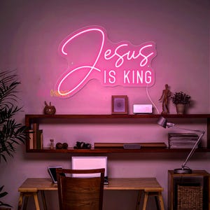 May include: Pink neon sign with the text "Jesus is King" on a white background. The sign is hanging on a wall in a room with a desk, chair, and other decor.