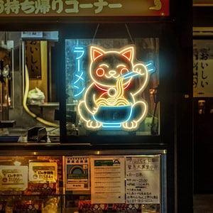 Puede incluir: Un letrero de ne&oacute;n de un gato de dibujos animados comiendo fideos ramen. El letrero est&aacute; en el escaparate de una tienda con otros letreros que anuncian comida para llevar y ofertas de trabajo. El letrero dice "Ramen" en caracteres japoneses.