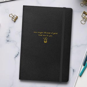 Puede incluir: Diario negro con el texto dorado "one single thread of gold tied me to you". El diario tiene un cierre de banda elástica y una ilustración dorada de manos entrelazadas.