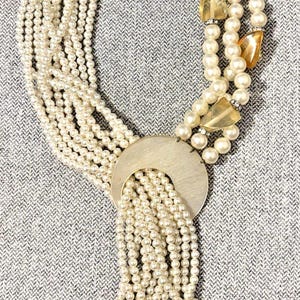 May include: A multi-strand necklace featuring numerous small white faux pearls arranged in a braided pattern. A large, circular silver-toned metal pendant acts as a focal point, with several strands of pearls cascading downward from its center.
