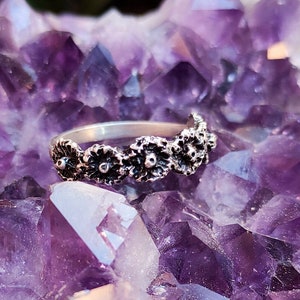 May include: A silver ring featuring a floral design. The ring has a band of small, detailed flowers. The ring is displayed on a background of purple amethyst crystals, creating a contrast of textures and colors.