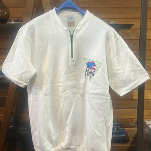 Peut inclure: Chemise à manches courtes blanche avec une fermeture éclair verte et un logo Puma sur le devant. Le logo est un triangle bleu et vert avec un logo Puma blanc à l'intérieur.