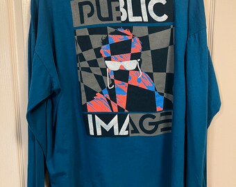 Può includere: Una maglietta a maniche lunghe blu turchese con una stampa grafica di una persona con occhiali da sole e il testo "PUBLIC IMAGE" in lettere bianche e grigie.