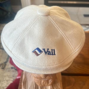 May include: Off-white newsboy cap with the word "Vail" embroidered in navy blue, with a blue and brown logo. The cap has a rounded top with a small button and stitched panels. The cap is made of a soft, textured material.